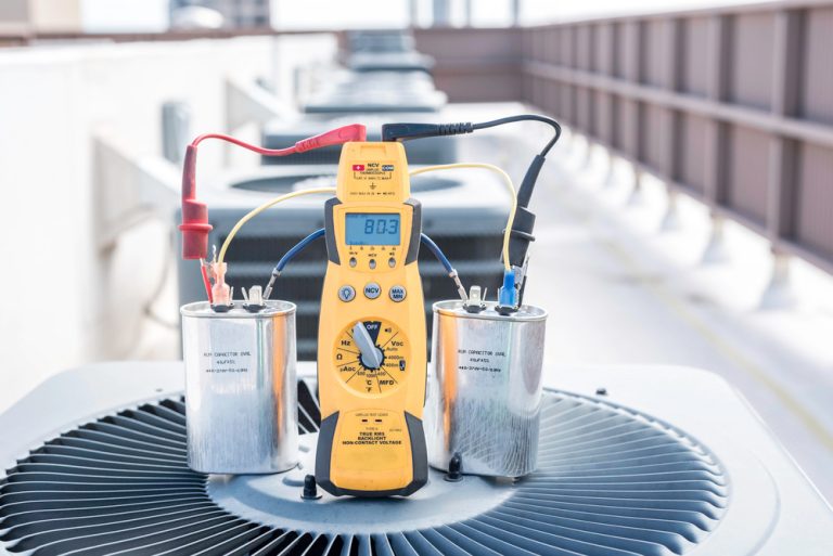 Technician using yellow multimeter to test microfarad reading on dual run capacitor during air conditioner replacement in Katy TX, atop outdoor HVAC unit.